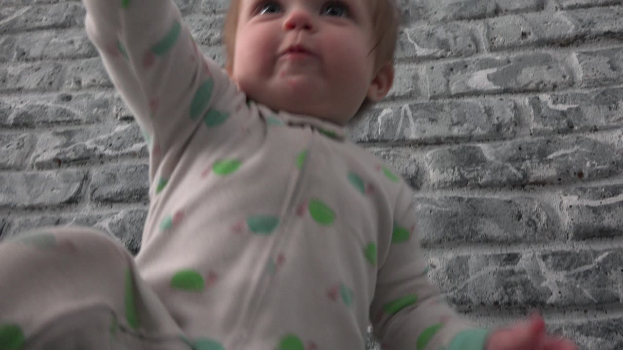 Toddler reaching upward near a gray brick-pattern wall.