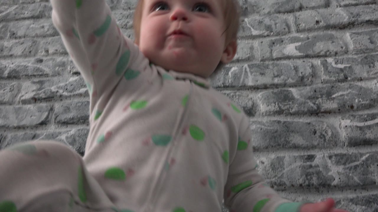The toddler remains close to the ground-level camera in Westminster, Colorado, looking upward with one arm lifted against the gray brick-pattern wall.