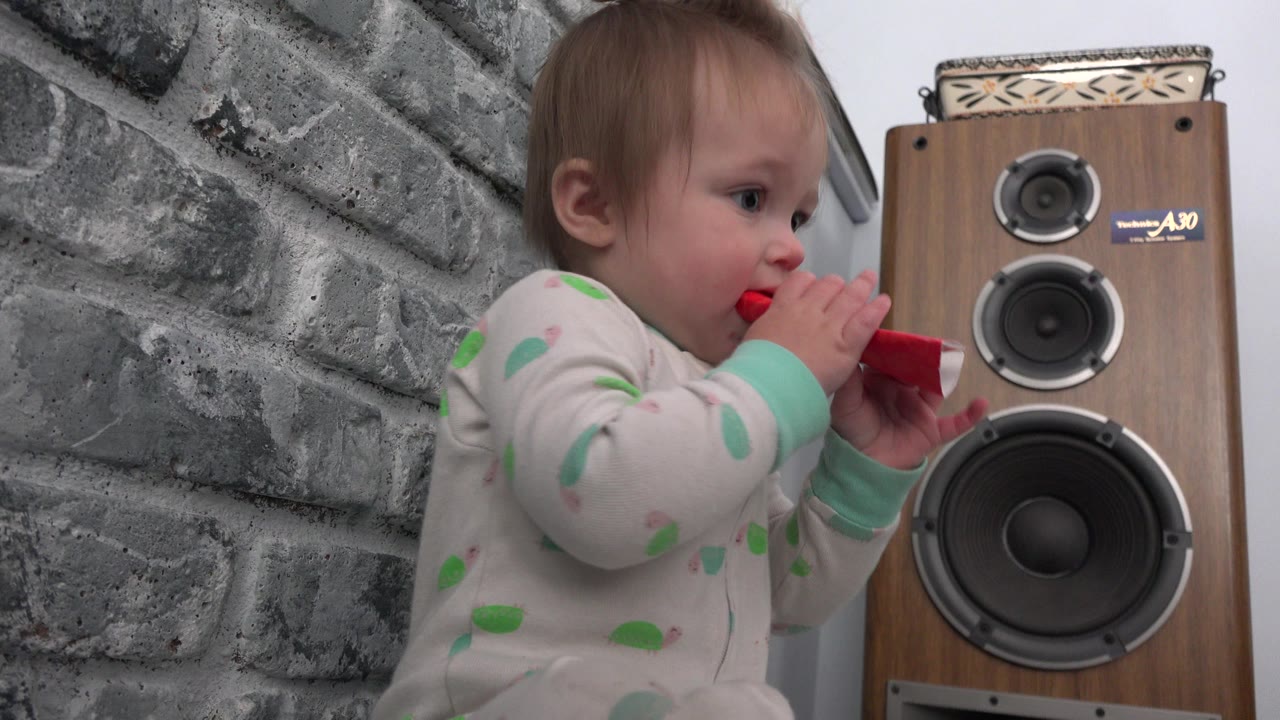 Baby holding a red snack tube beside a tall speaker.