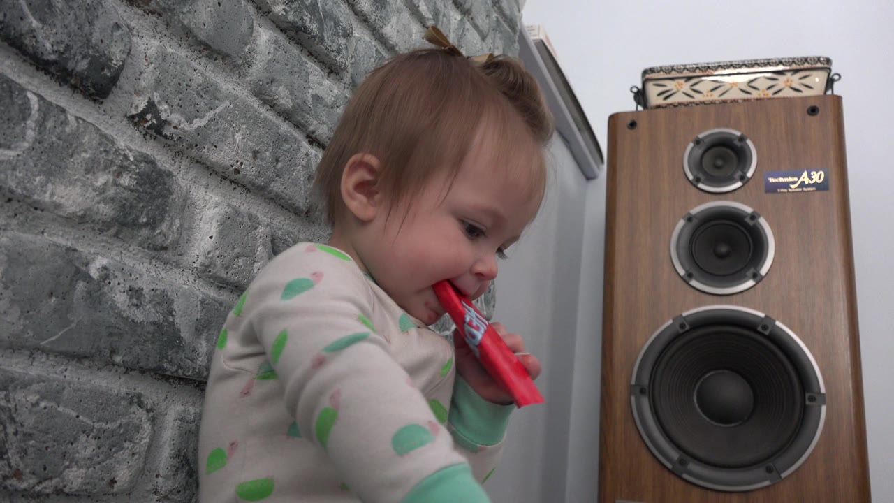 Baby holding a red package near a tall wooden speaker.