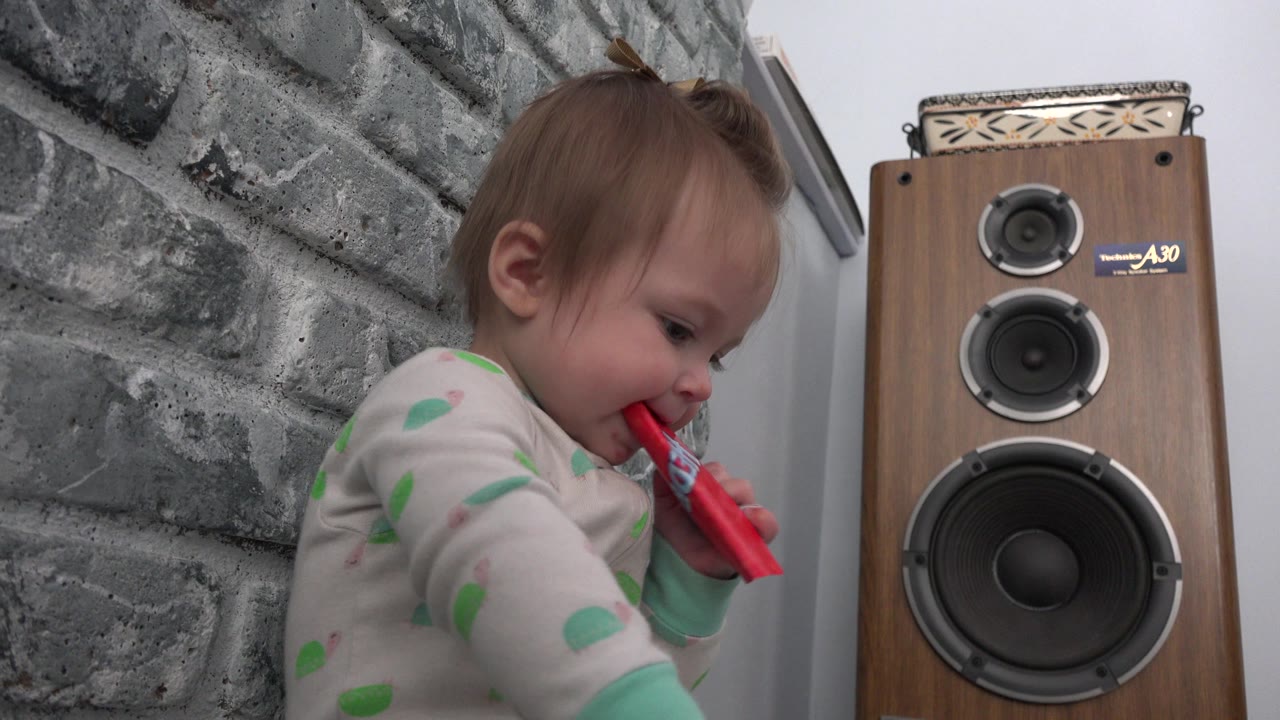 At 0:01 in Westminster, Colorado, the baby keeps the red package at the mouth while facing down near the large wood-grain speaker against the gray wall.