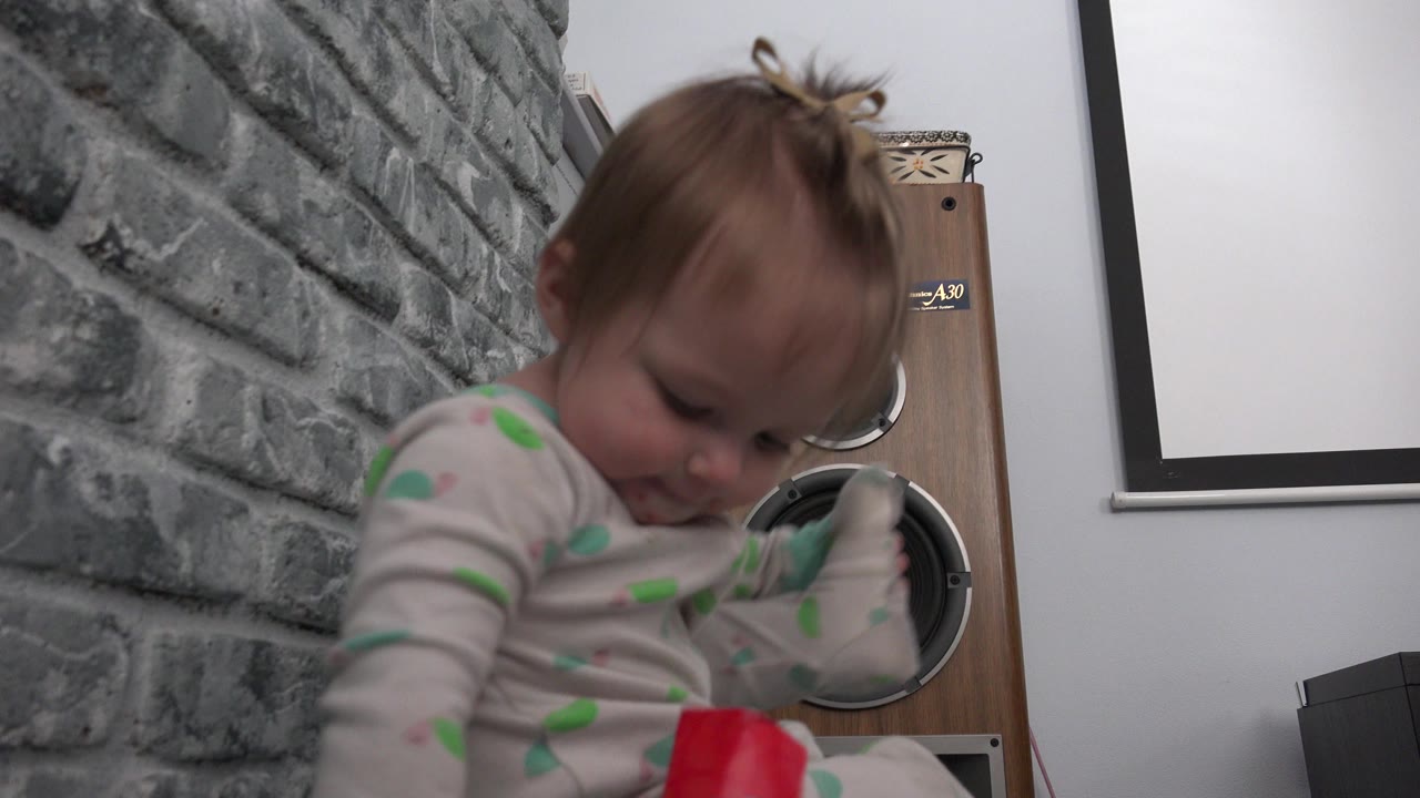 A young child in spotted pajamas sits on the floor in a Westminster, Colorado interior, looking down near a red paper or wrapper with a gray brick-textured wall behind them.
