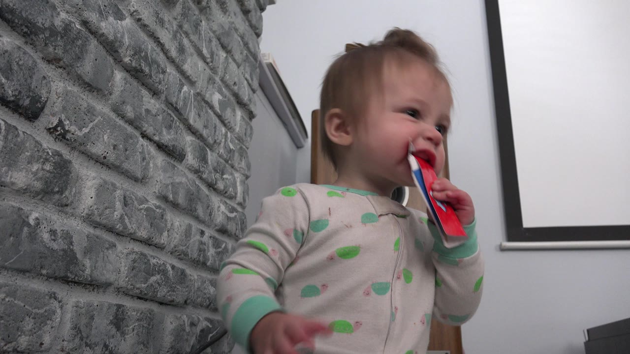 In Westminster, Colorado, the baby boy smiles with the red paper item in his mouth while seated beside the textured gray brick wall.