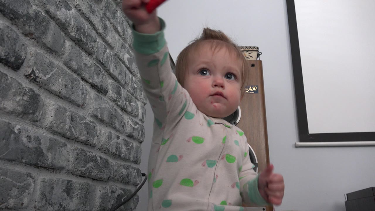 Baby reaches upward toward a red object near a gray brick-textured wall.