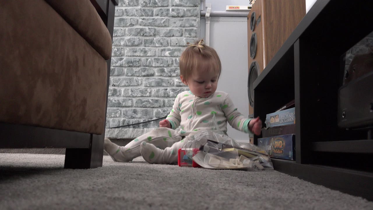In Westminster, Colorado, a blonde toddler sits on gray carpet beside a clear plastic bag, with a sofa at left and a dark media shelf at right.