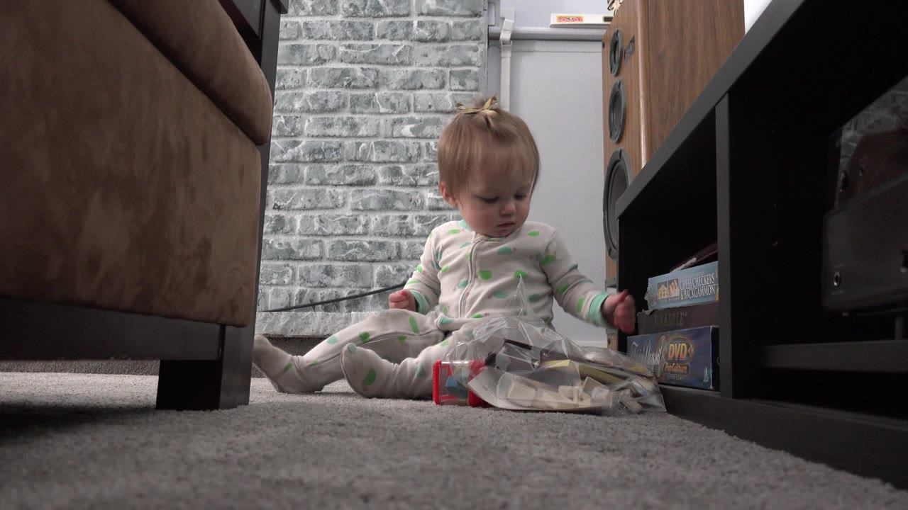 In Westminster, Colorado, the toddler looks down toward the clear bag on the carpet, seated between the sofa and a shelf holding game or DVD boxes.