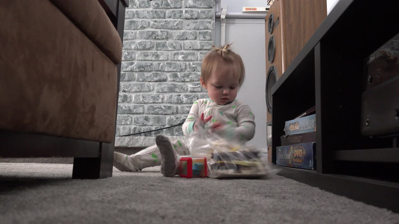 Baby in light pajamas sits on gray carpet in a Westminster, Colorado home interior, handling a clear plastic bag beside a red block near a sofa and shelving unit.