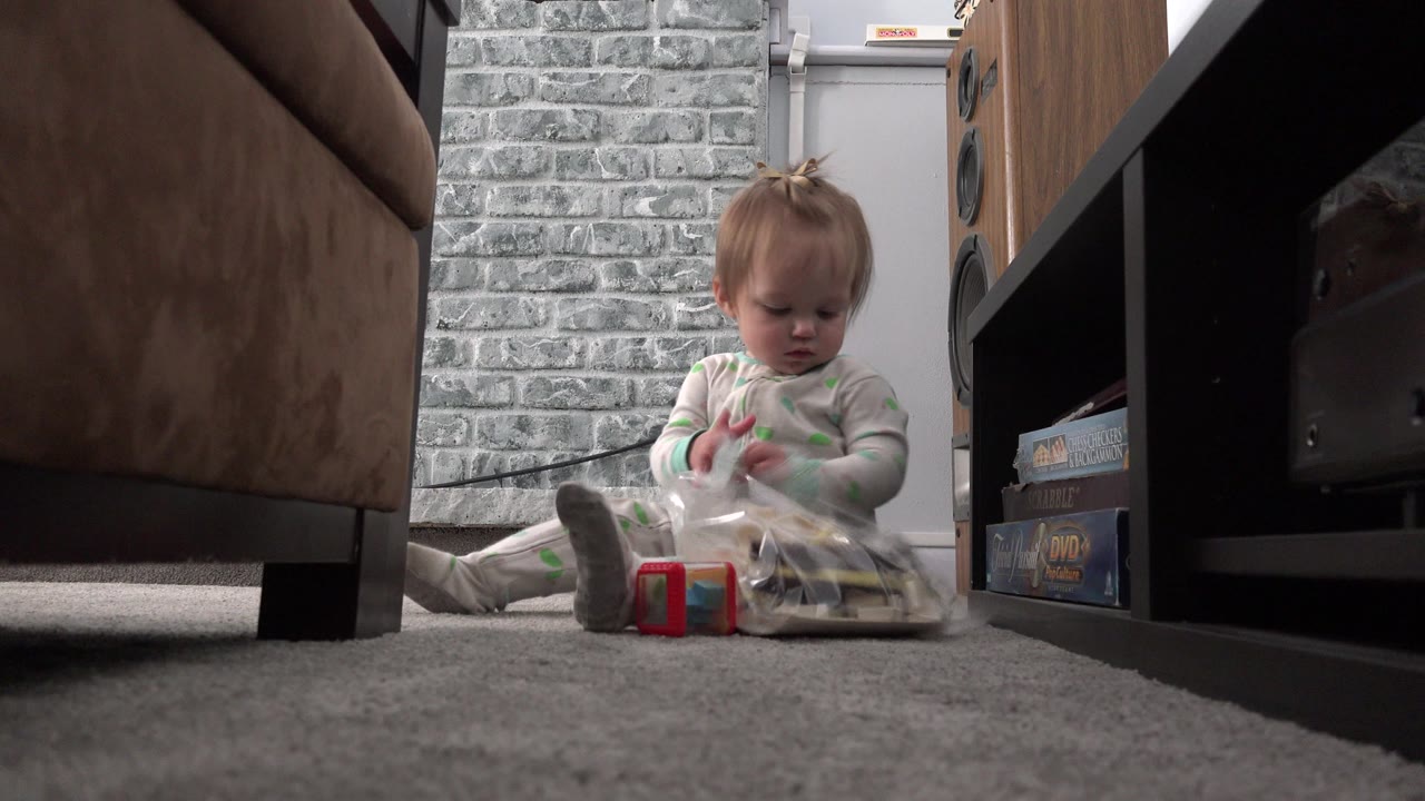 Baby in pajamas looks down at a clear plastic bag on the carpet in the Westminster, Colorado living room, with a red block in front and shelving to the right.