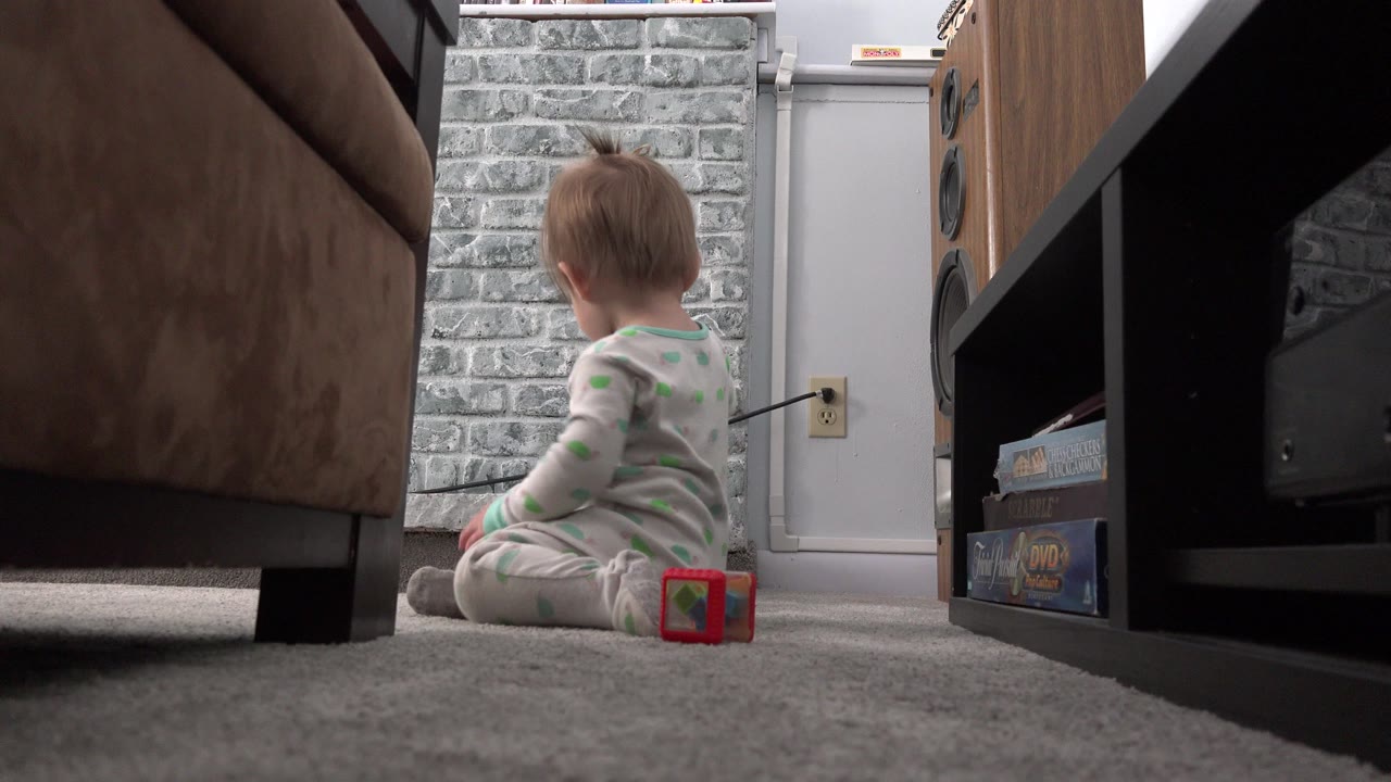 At 0:01, the baby remains seated with their back turned, positioned between a brown sofa and a black media shelf in the Westminster, Colorado living room.
