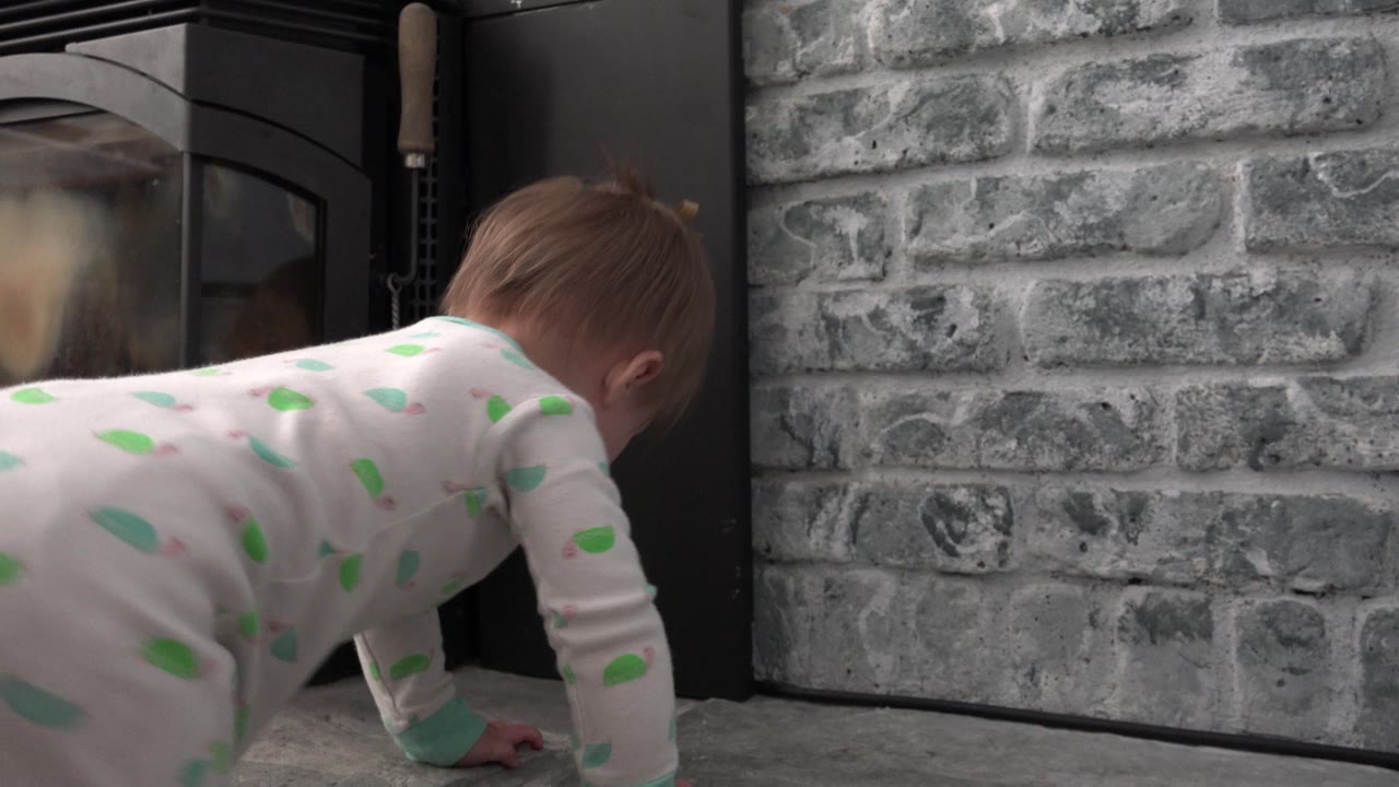 A blonde baby girl crawls low across a gray hearth floor beside a black fireplace and gray brick wall in Westminster, Colorado.