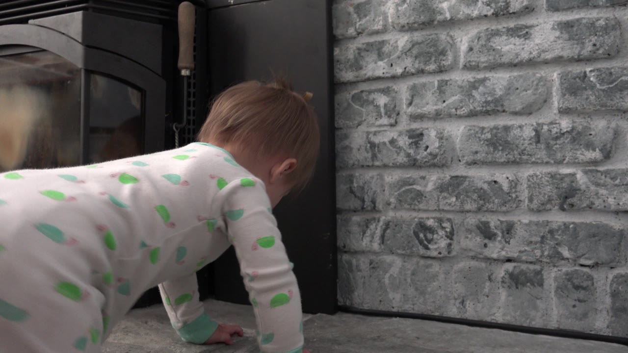 The blonde baby girl continues crawling near the black fireplace door, with the gray brick wall filling the background in Westminster, Colorado.