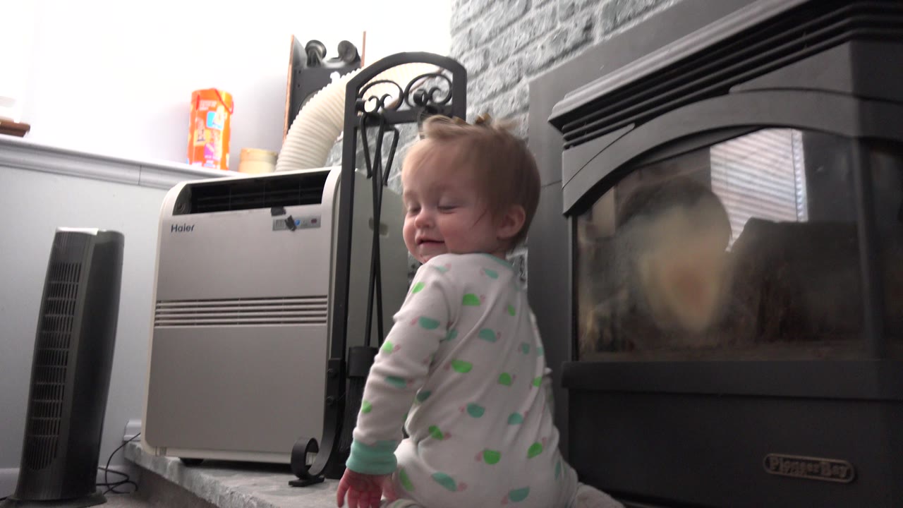 A baby in white and green pajamas stands near a black glass fireplace in a Westminster, Colorado home, with a portable air conditioner and fireplace tools behind her.