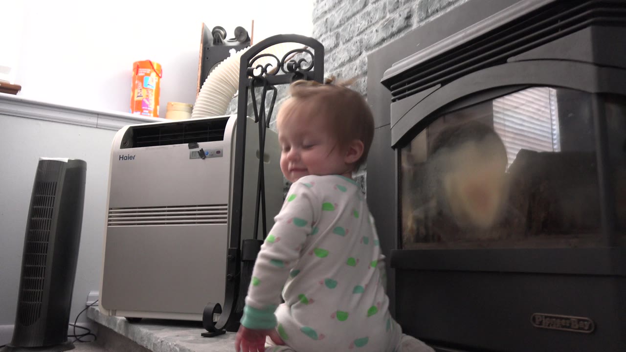 The baby smiles while standing beside the fireplace, with the glass door, gray hearth, and portable air conditioner visible in the Westminster, Colorado home interior.