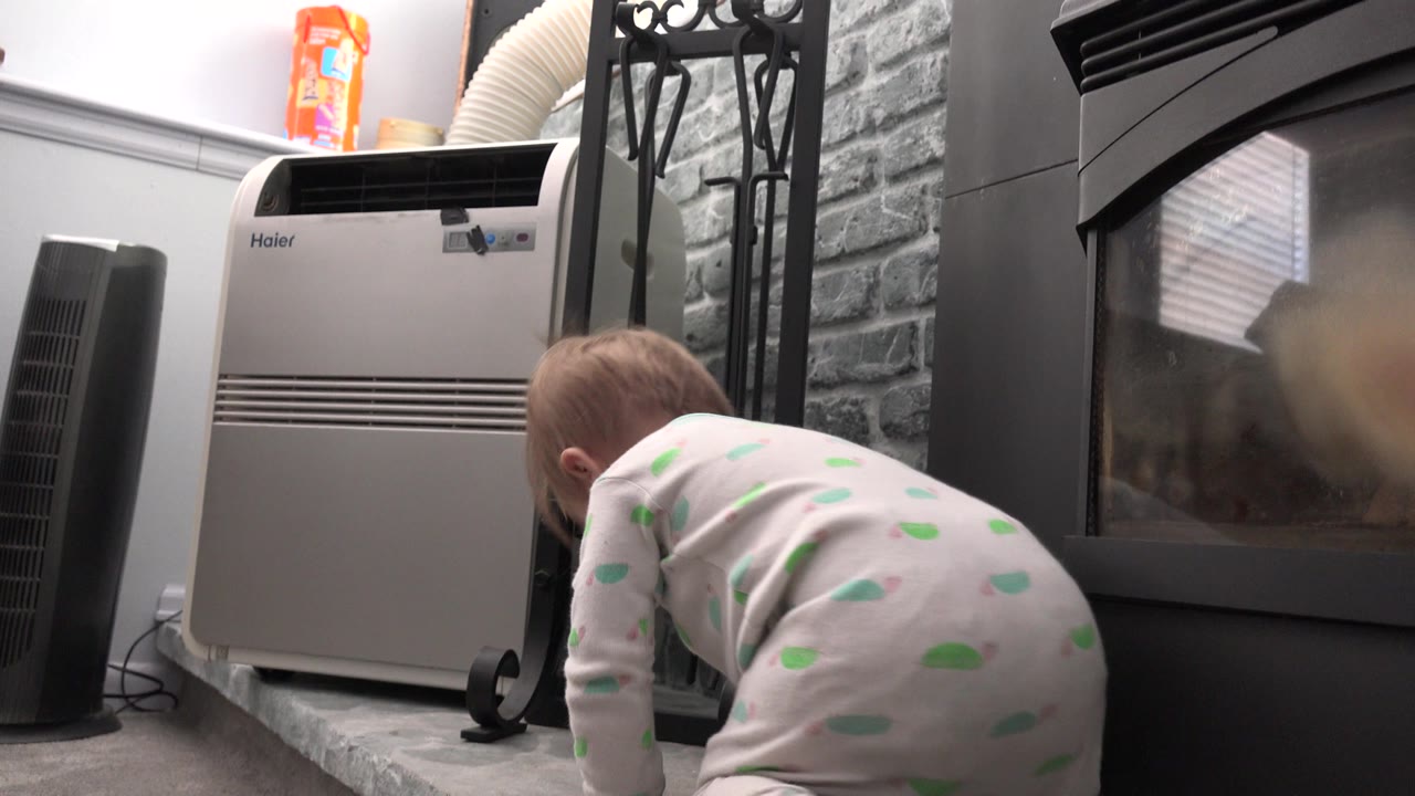 Baby crawling on a stone fireplace hearth beside a black fireplace and household appliance.