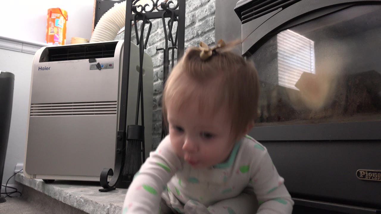 Baby leaning forward on a low hearth beside a black stove or heater.