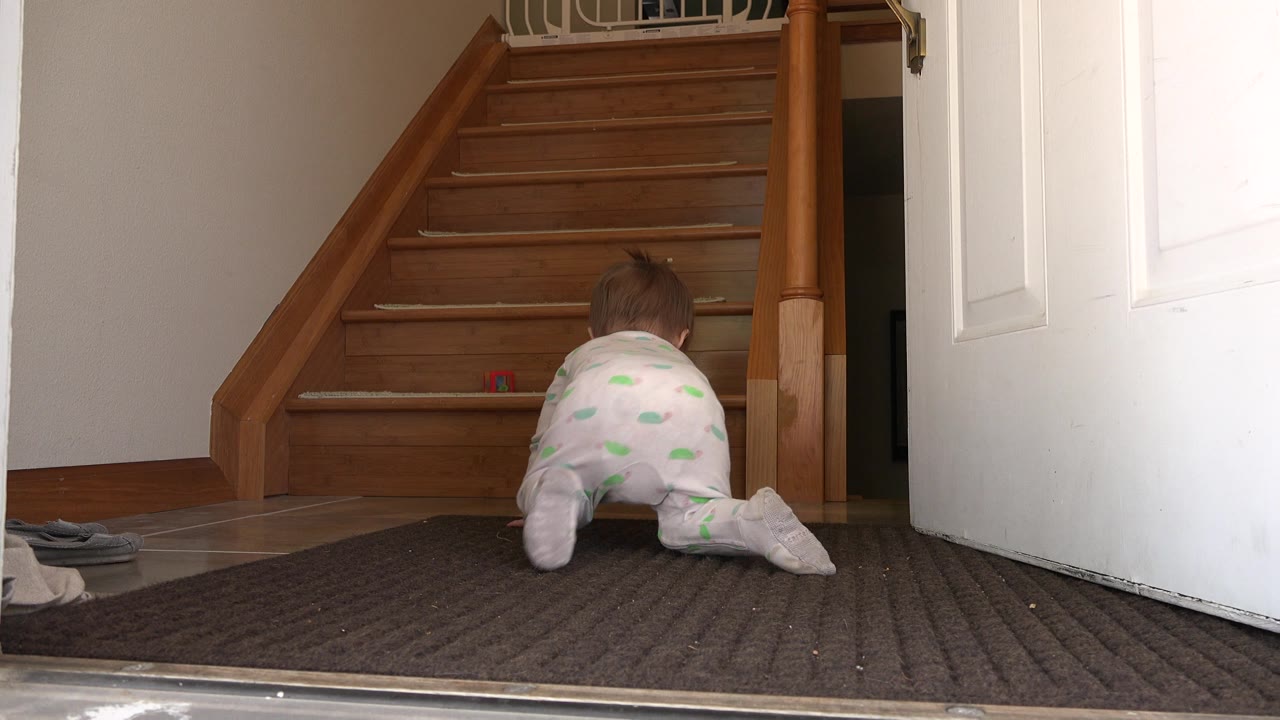 In a Westminster, Colorado home interior, a baby in pale pajamas crawls away from the low camera toward wooden stairs, with an open white door and dark mat in the entryway.
