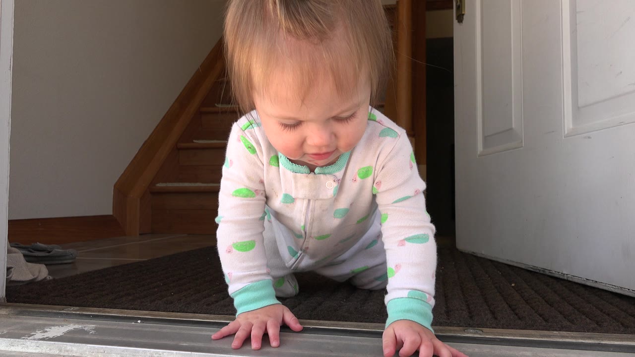 A baby in a pale sleeper crawls on a dark entry carpet at a Westminster, Colorado home doorway, with the child's head tilted down and a wood staircase behind.