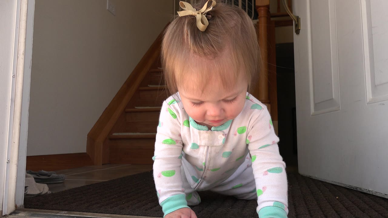 At 0:00 in Westminster, Colorado, a baby girl in a light sleeper crawls toward the low camera on a dark mat beside an open white doorway, with a wooden staircase behind her.