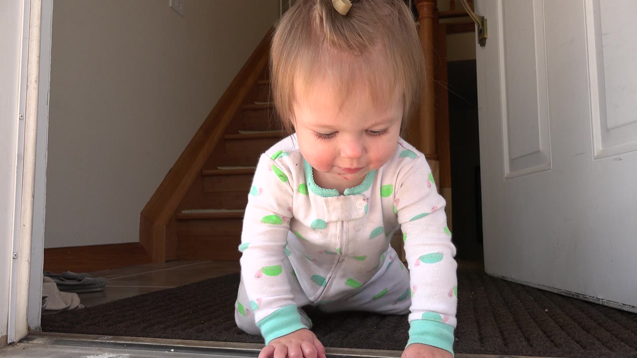 The baby remains on hands and knees near the doorway, looking down while the staircase and open door frame the Westminster home interior.