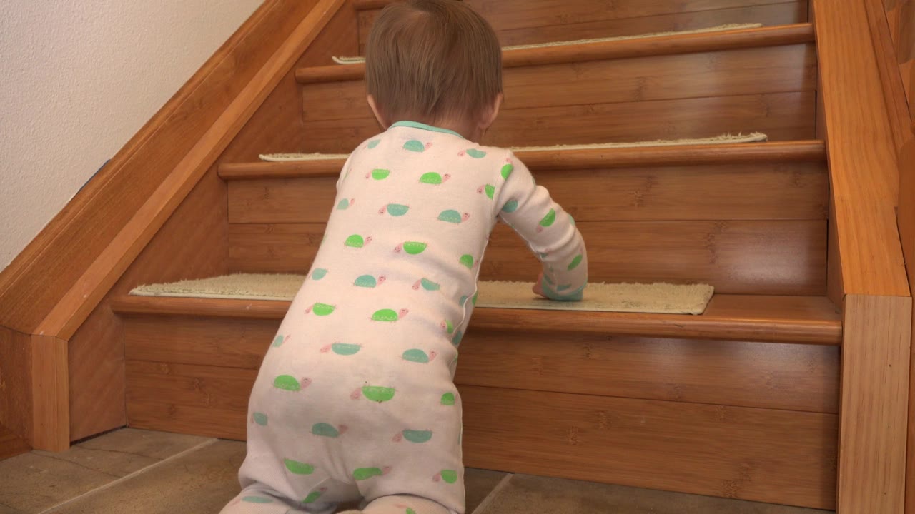 The toddler leans farther onto the first step, with the polished wooden stairs and beige tread mats filling the frame.