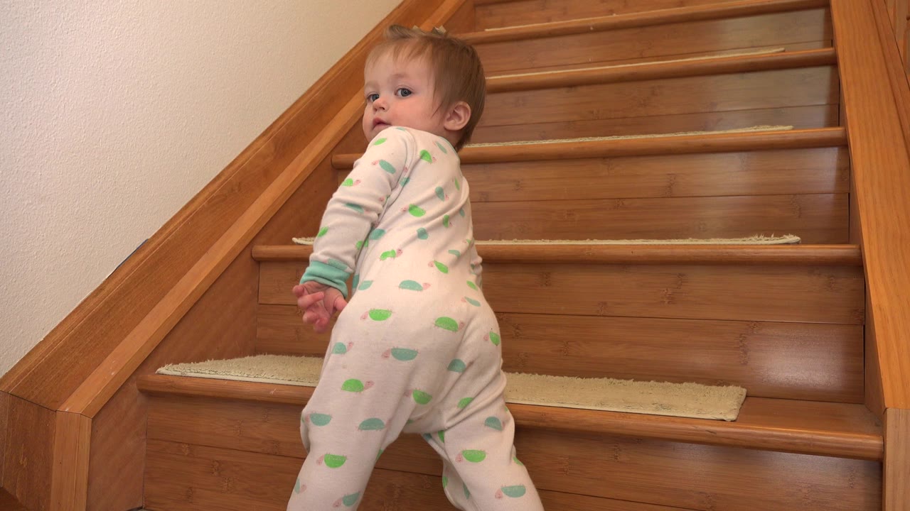 Infant in a green and white sleeper stands near wooden stairs.
