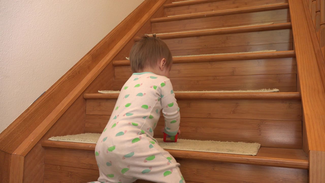 At 0:01, the baby continues climbing the wooden steps, seen mostly from behind with a red and green toy near her hand.