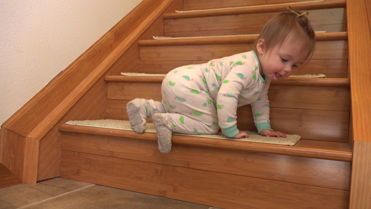In Westminster, Colorado, a baby girl in a white and green onesie crawls on a lower wooden stair with beige tread pads and a wood railing behind her.