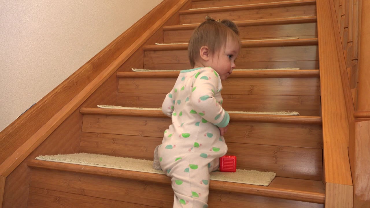 An infant girl in white green-patterned pajamas kneels on a wooden staircase in Westminster, Colorado, with a small red block beside her on the step.