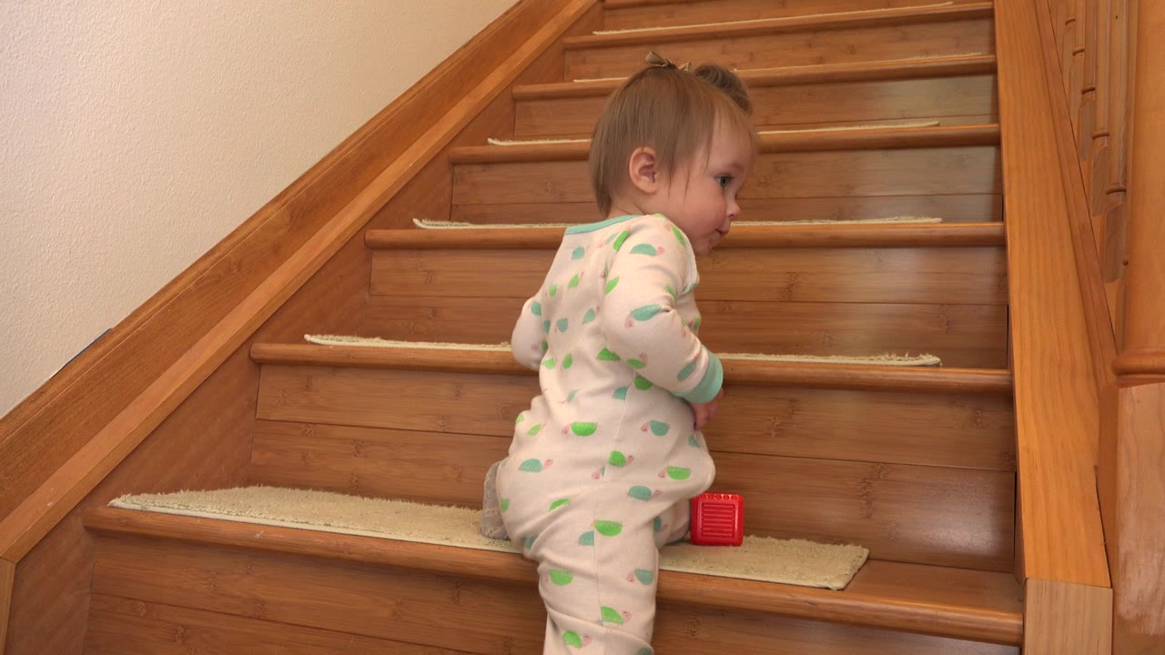 The infant girl faces up the wooden staircase in Westminster, Colorado, with her hands near the next step and the red block tucked beside her knee.
