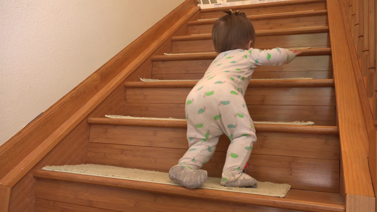 A baby in pale patterned footed pajamas climbs the lower wooden stairs inside a Westminster, Colorado home, seen from behind near the railing.