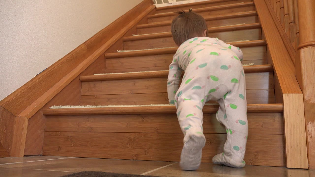 A baby in white footed pajamas with green patterns climbs the lower wooden stairs inside a Westminster, Colorado home, seen from behind near the bottom step.