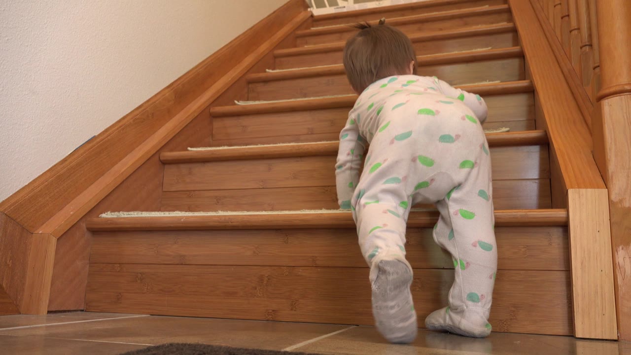 The baby continues crawling upward on the wooden staircase, with one socked foot lifted and the stair railings framing the climb in the Westminster home.