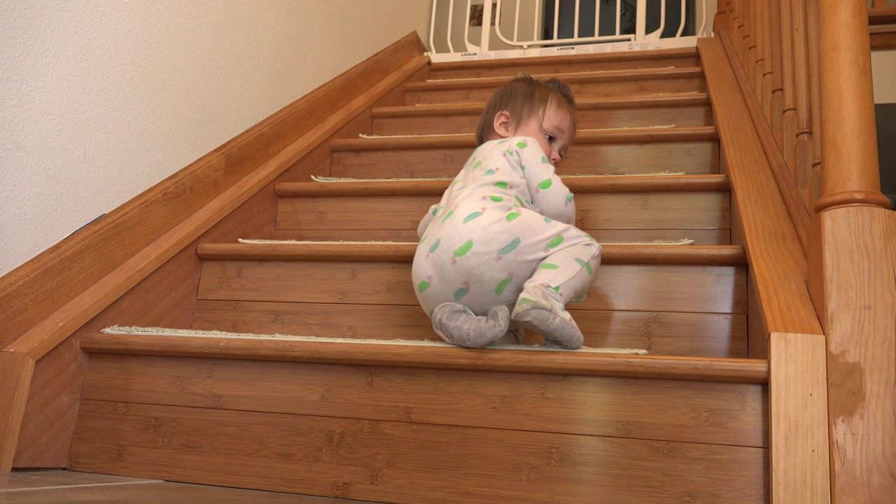 A baby in a white patterned sleeper sits on wooden stairs in a Westminster, Colorado home, turned partly toward the camera near the lower steps.
