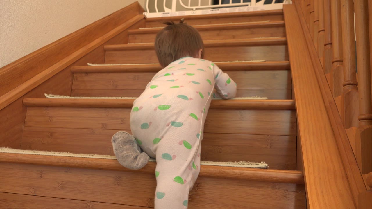 The child crawls one step higher on the wooden staircase, with gray pajama feet near the lower tread and the white gate visible above.