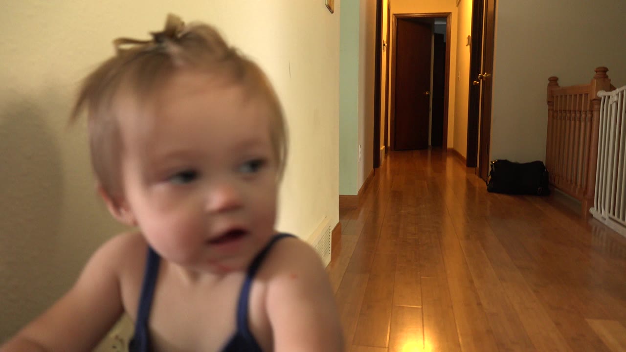 The toddler remains near the left foreground while the Westminster home hallway, wooden floor, open doors, and white baby gate stay visible in the background.