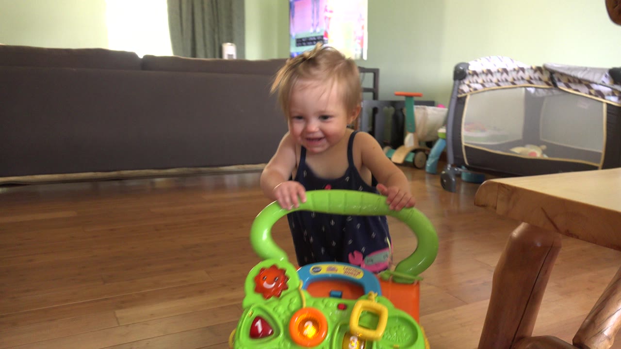 Toddler smiling behind a green toy walker in a living room.