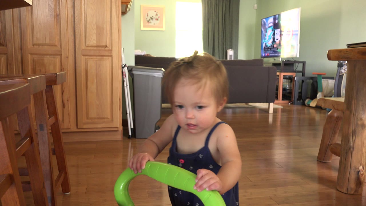 At 0:00, a baby stands low in frame behind a bright green toy handle inside a Westminster, Colorado home, with wood cabinets and chairs around the room.