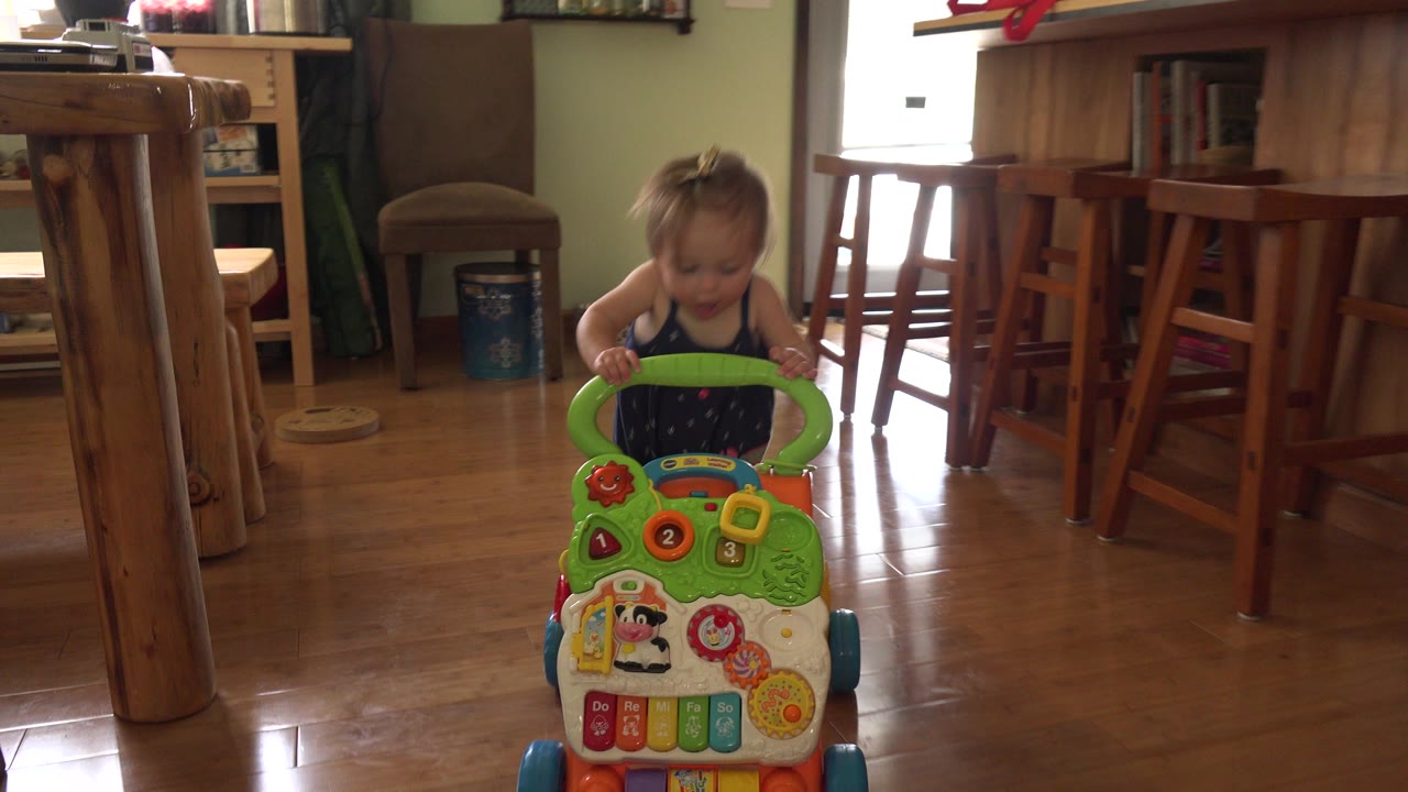A blonde baby in Westminster, Colorado holds a green activity walker near the lower center of the frame, with wooden furniture and bright window light behind.