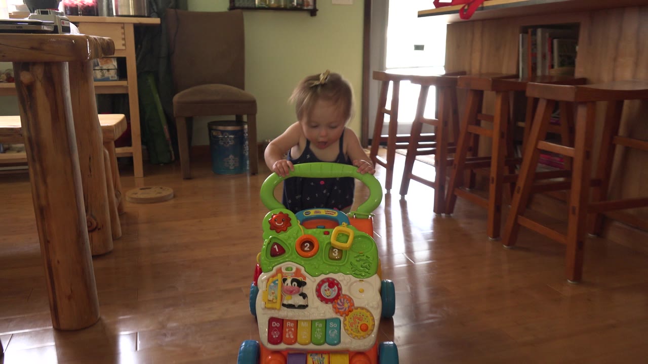 The baby looks down at the green handle of the activity walker, with the wooden floor reflecting daylight and a kitchen island in the background.