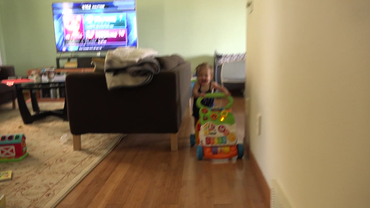 A young child grips the green handle of a toy walker at the right edge of the frame, with the living room couch, blanket, coffee table, and television behind them.