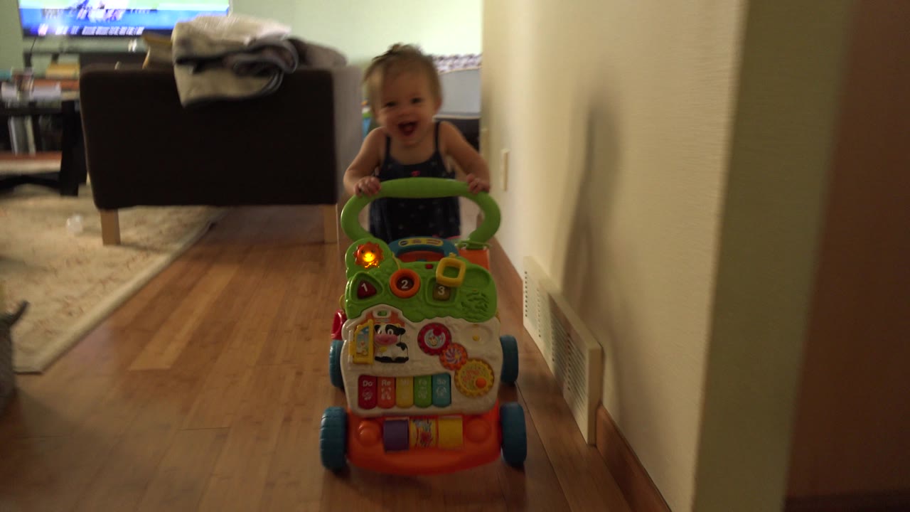 A smiling baby pushes a green activity walker with orange lights along the hallway, with the living room couch and television still behind the child.