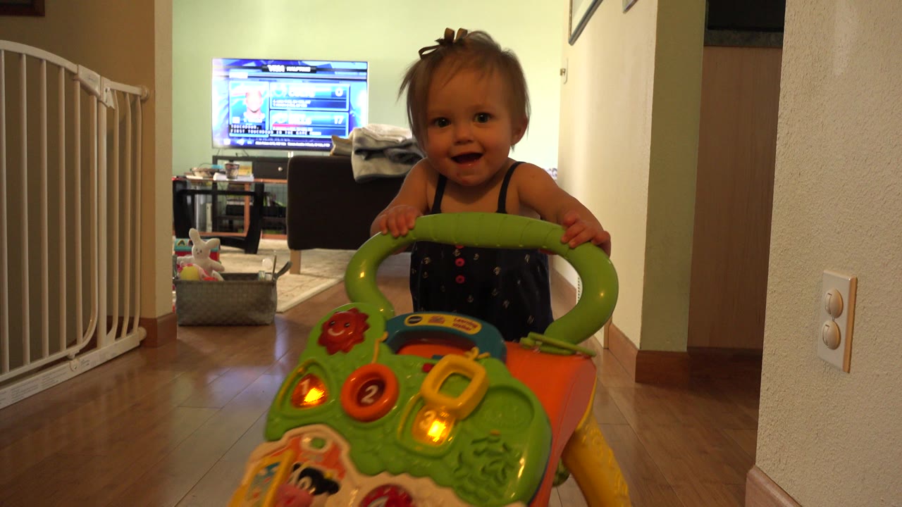 The toddler in Westminster, Colorado smiles toward the low camera while gripping the green handle of a push toy in the hallway.