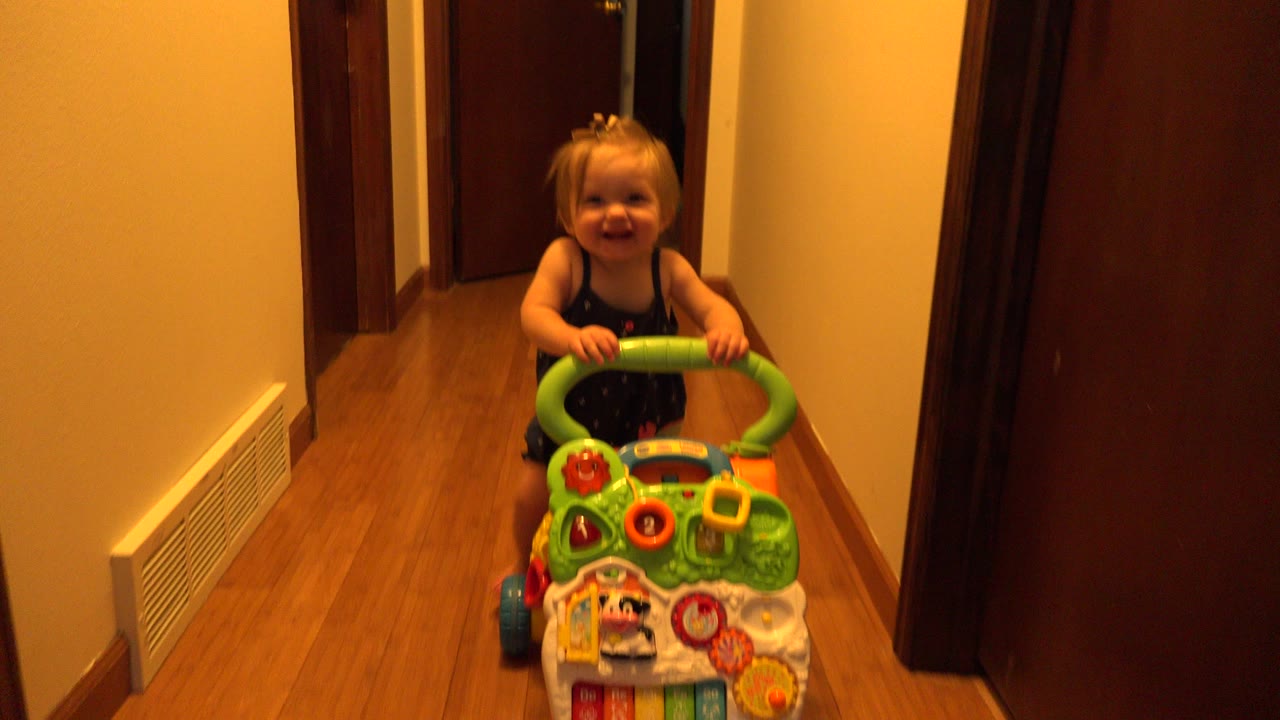 Toddler smiling behind a green toy cart in a hallway.