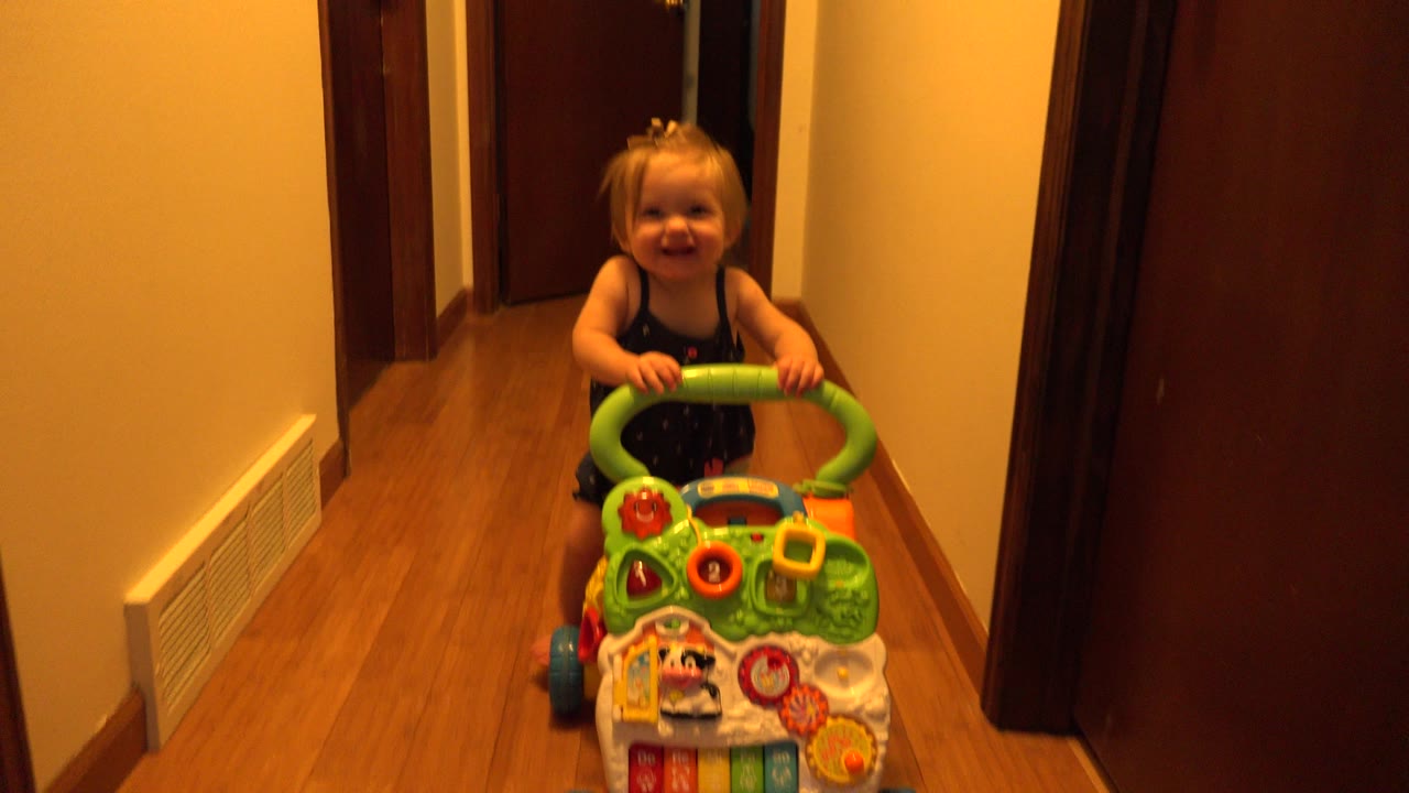 The toddler stands behind the green toy cart in the narrow Westminster, Colorado hallway, with doors and a wall vent visible around her.