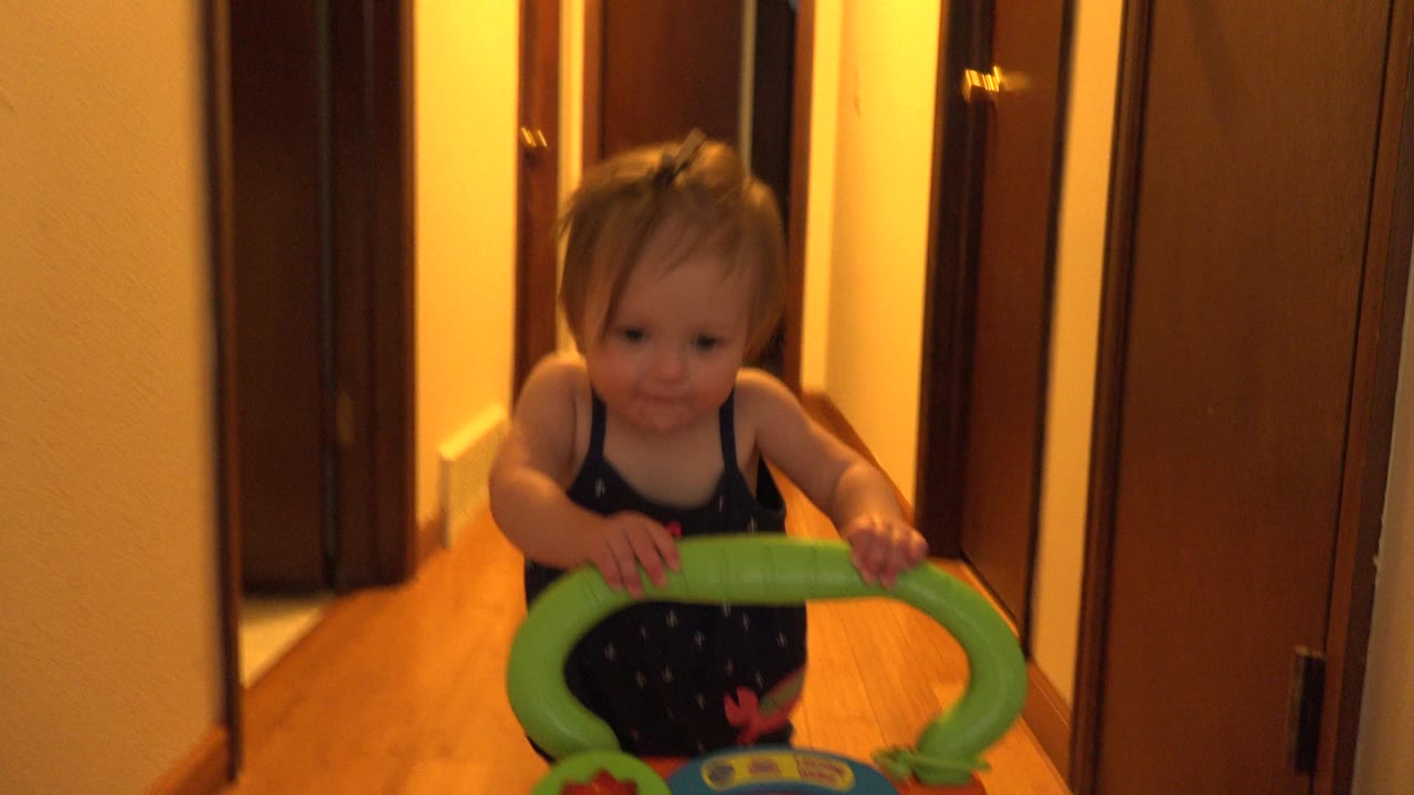 A baby in Westminster, Colorado holds a green toy walker while facing the camera in a warm-lit hallway with wood flooring and brown doors.