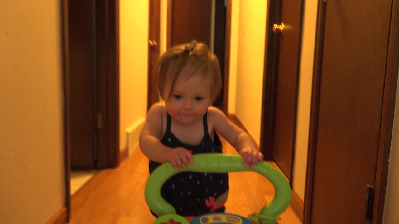 The baby remains centered behind the green walker in the Westminster home hallway, with warm light reflecting on the wood floor and doors along both sides.
