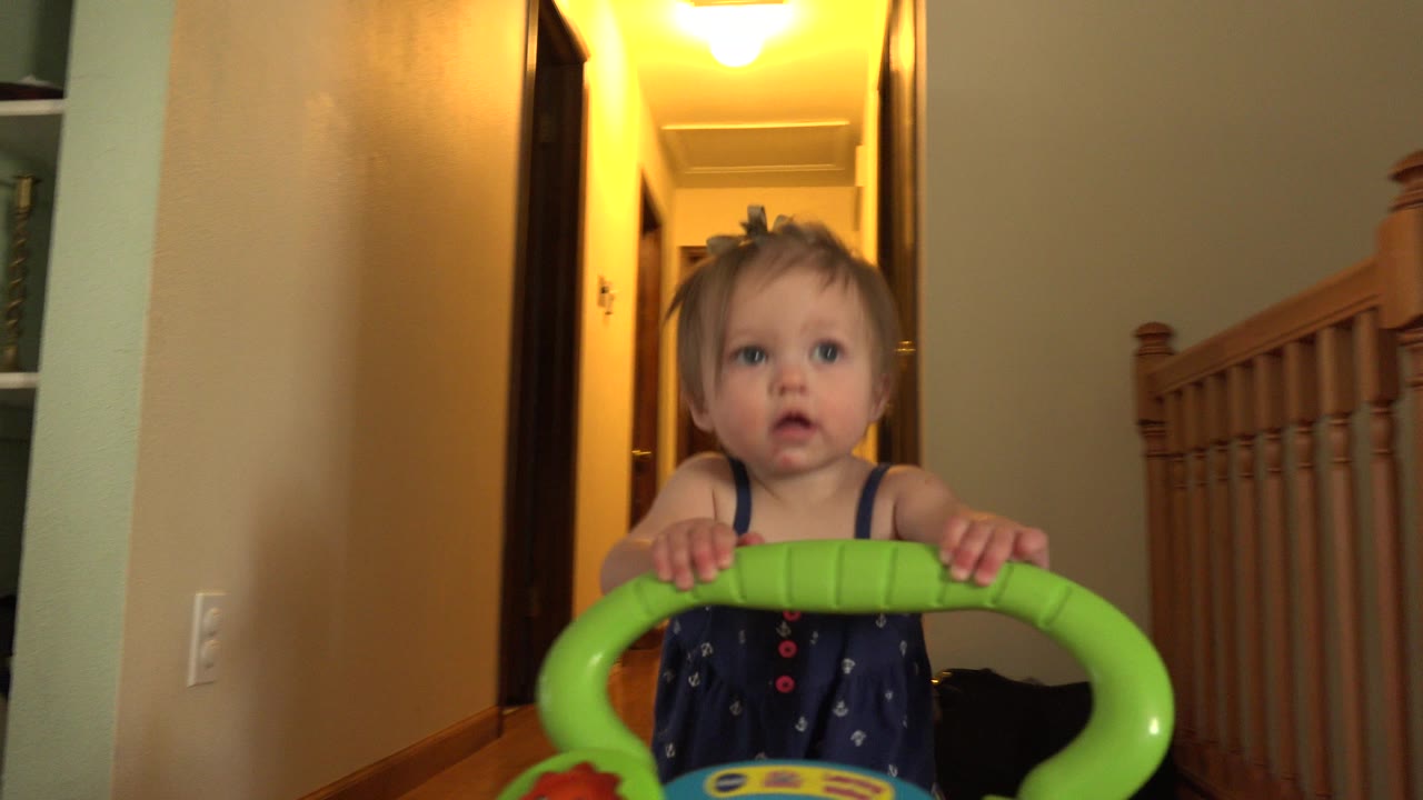 The child remains centered behind the green walker, with the hallway light glowing overhead and the stair railing visible along the right side of the Westminster home interior.