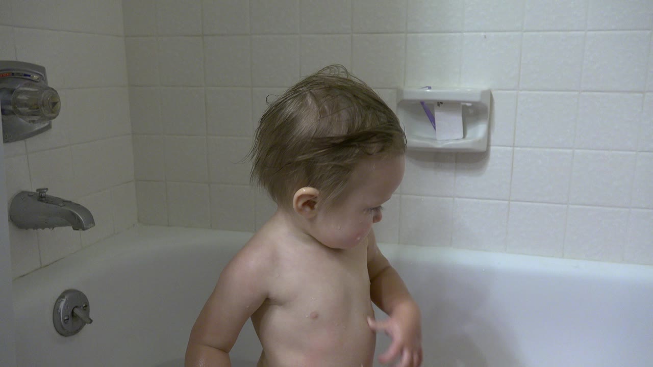 A toddler stands in a white bathtub in Westminster, Colorado, looking down with a chrome faucet and white tiled wall behind him.