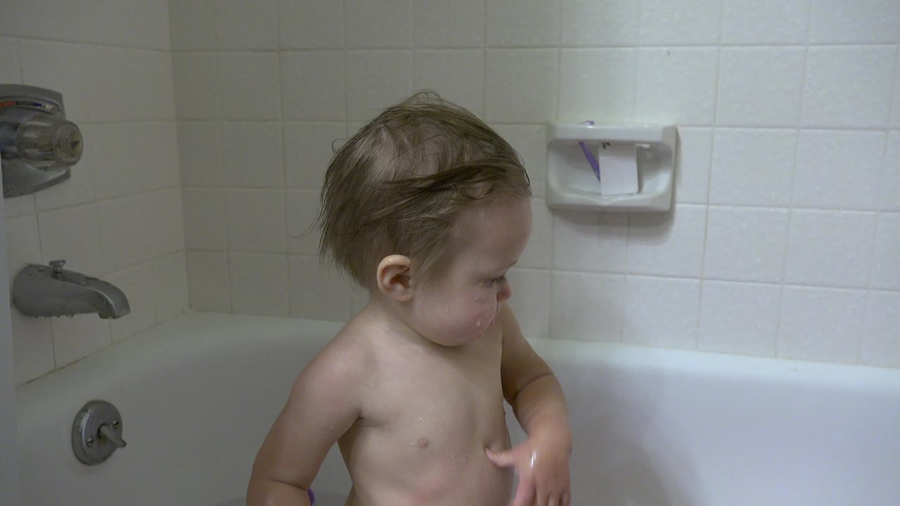 Toddler standing in bathtub with one hand raised near his chest.