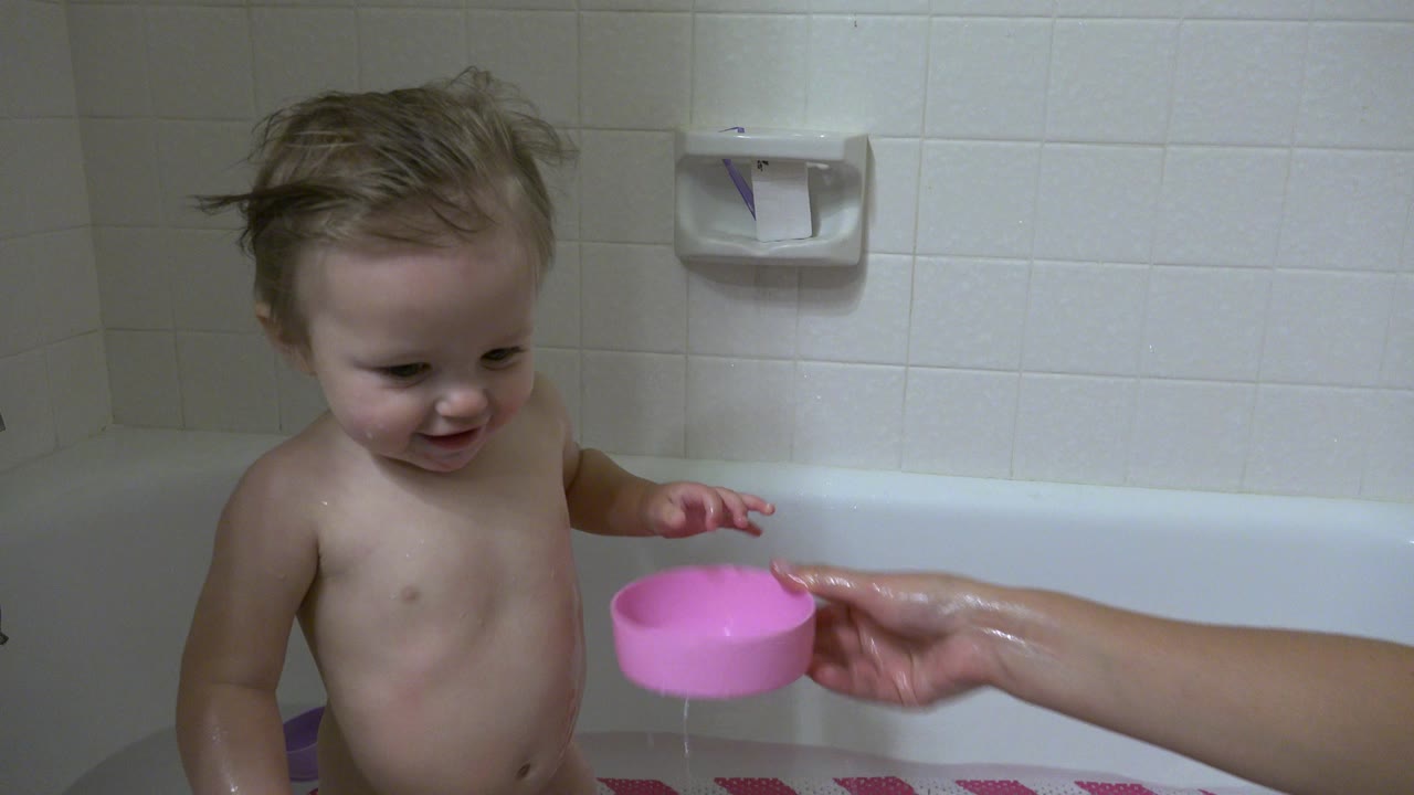 Young child in a white bathtub beside a pink bowl held by an adult hand.